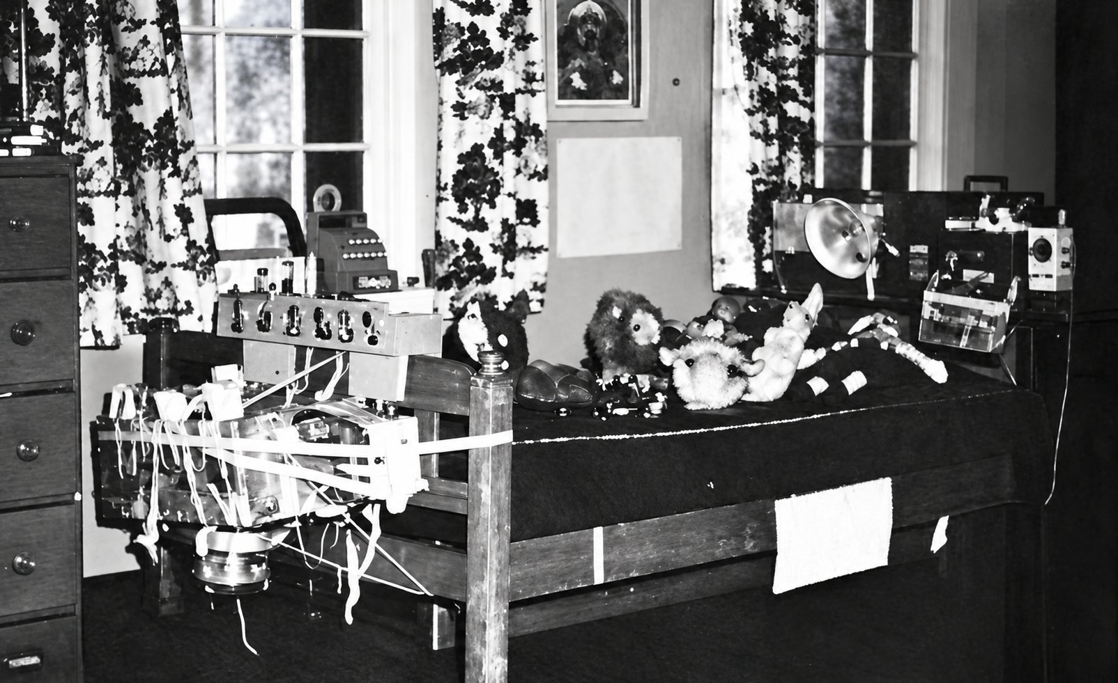Photo ancienne en noir et blanc d'un comptoir de boutique sur lequel sont posés une caisse enregistreuse, des jouets et divers objets, devant une fenêtre aux rideaux fleuris.